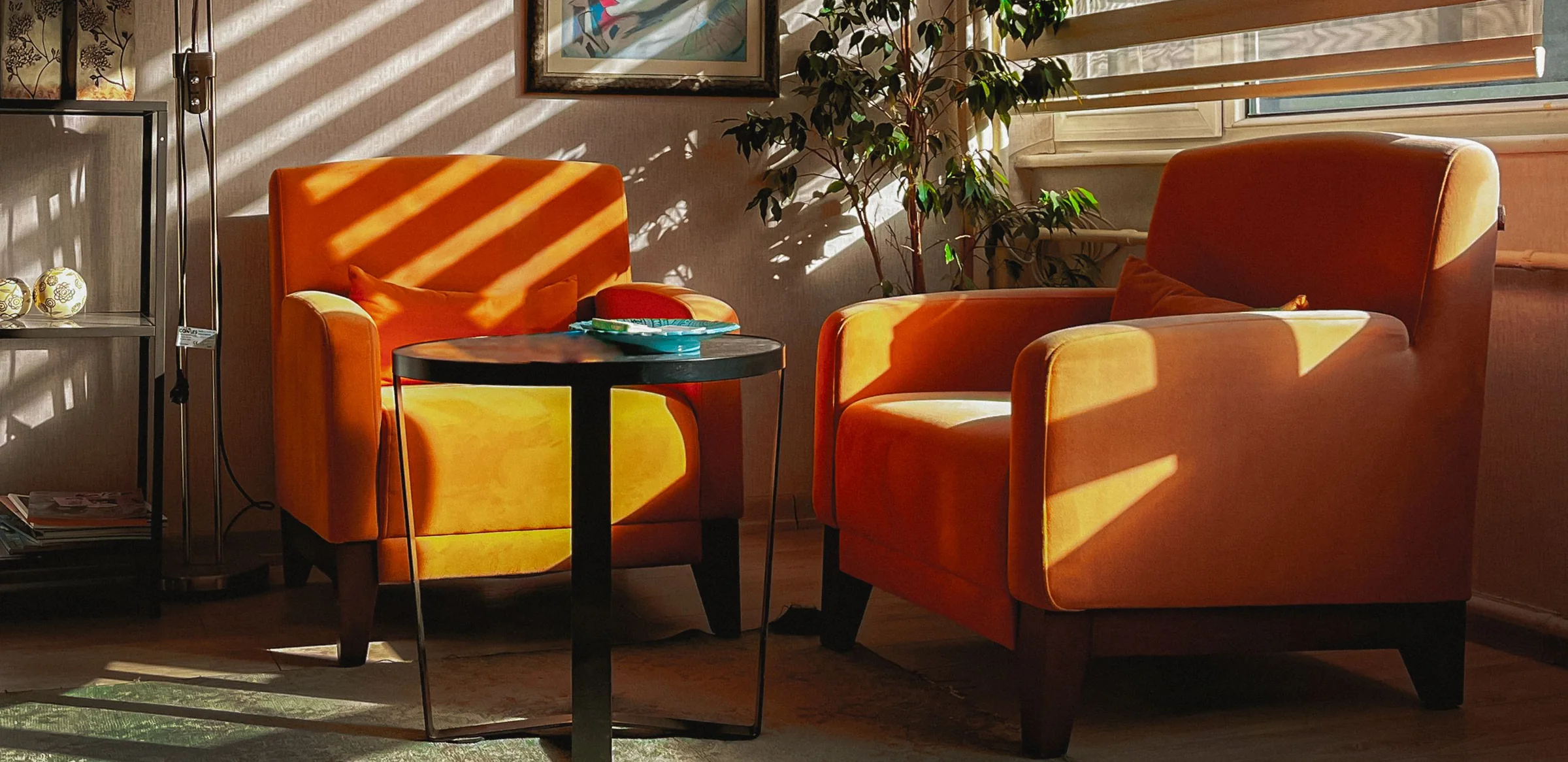 Deux fauteuils orange de part et d’autre d’une table basse, zébrés par le soleil qui passe à travers un store.