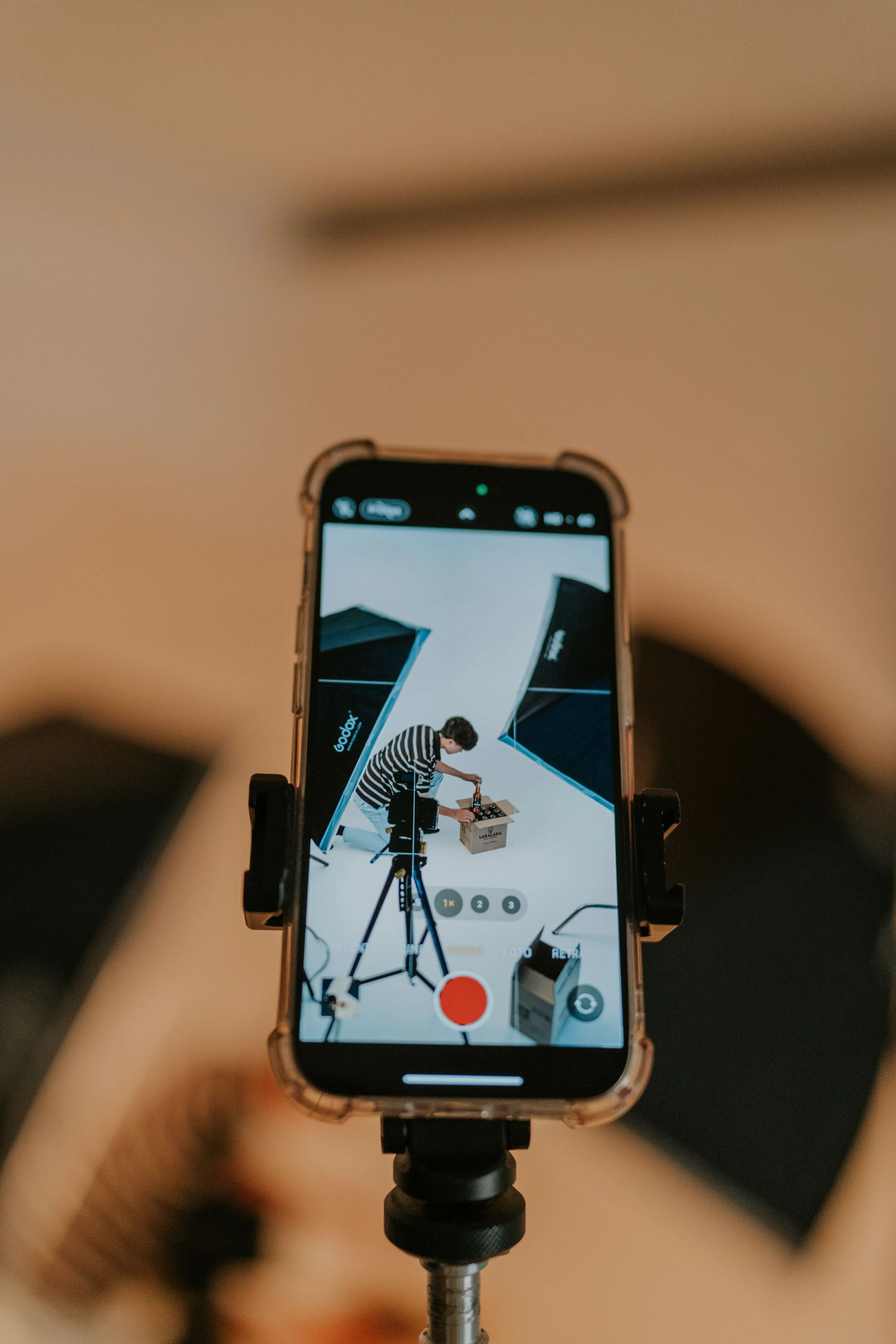 Un smartphone sur trépied filme un homme qui déballe un colis dans un studio aux tons chauds.
