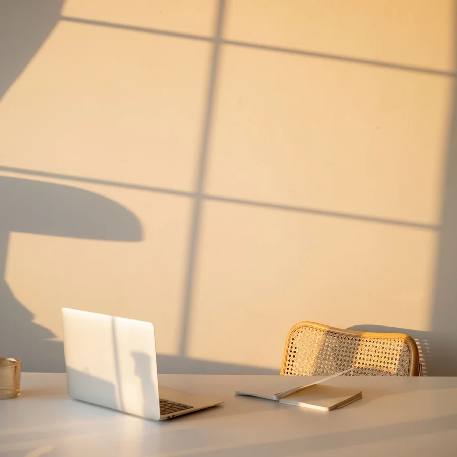 Un ordinateur portable ouvert et un carnet posés sur un bureau clair, devant un mur crème où le soleil de fin de journée dessine la grille d’une fenêtre.