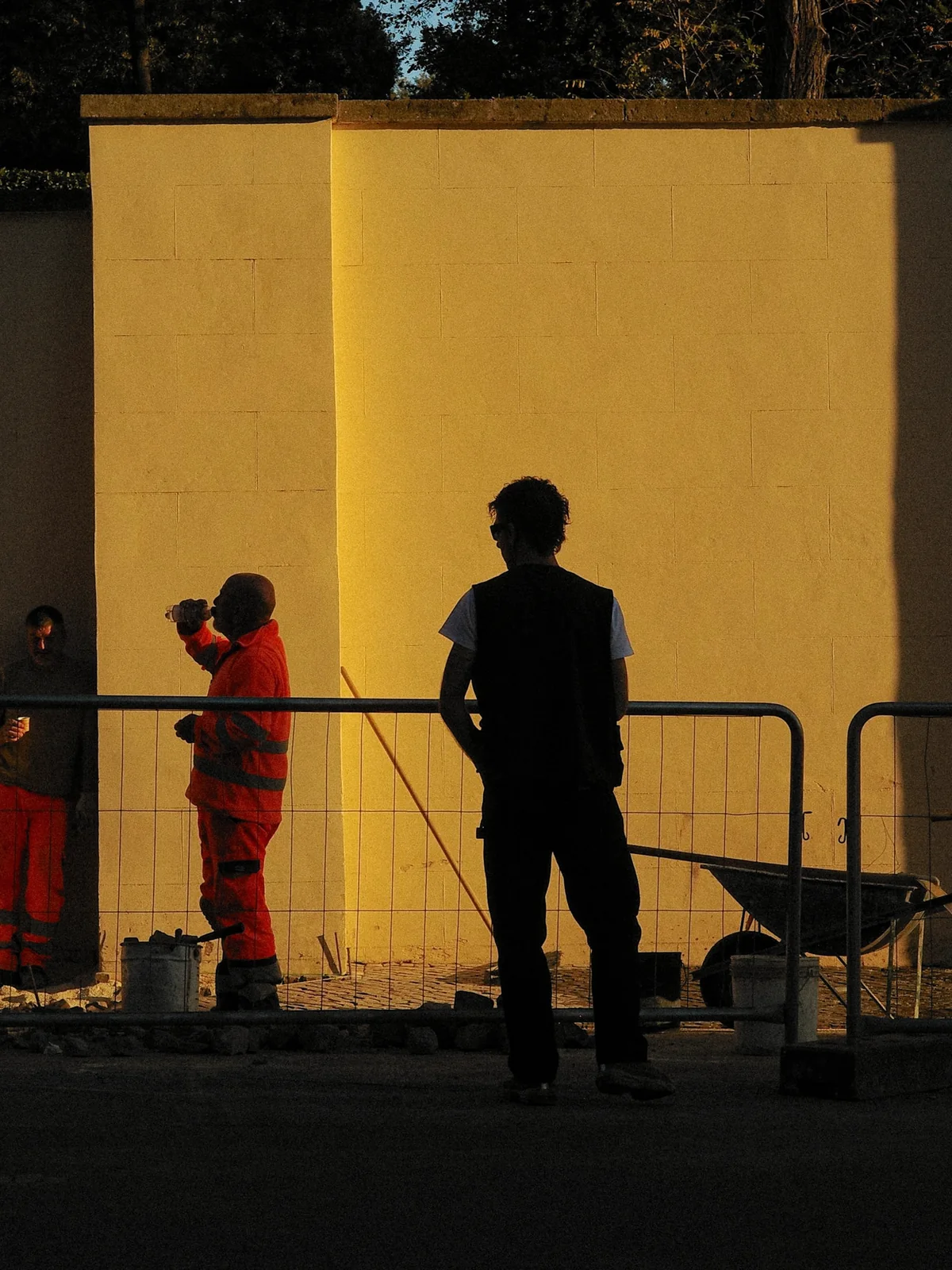 Trois ouvriers devant un long mur ocre en plein soleil rasant de fin de journée, derrière une barrière de chantier, avec une brouette, des seaux et des pavés en cours de pose.