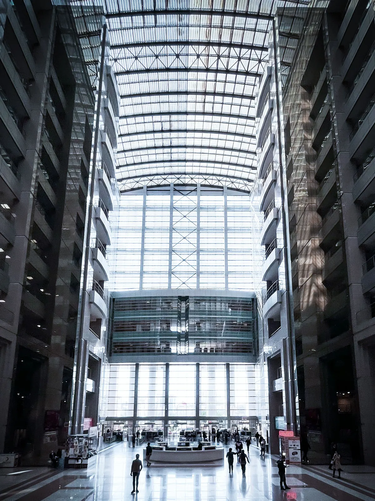 Le hall vitré d’un immeuble de bureaux, quelques silhouettes traversant le dallage poli sous une haute verrière.