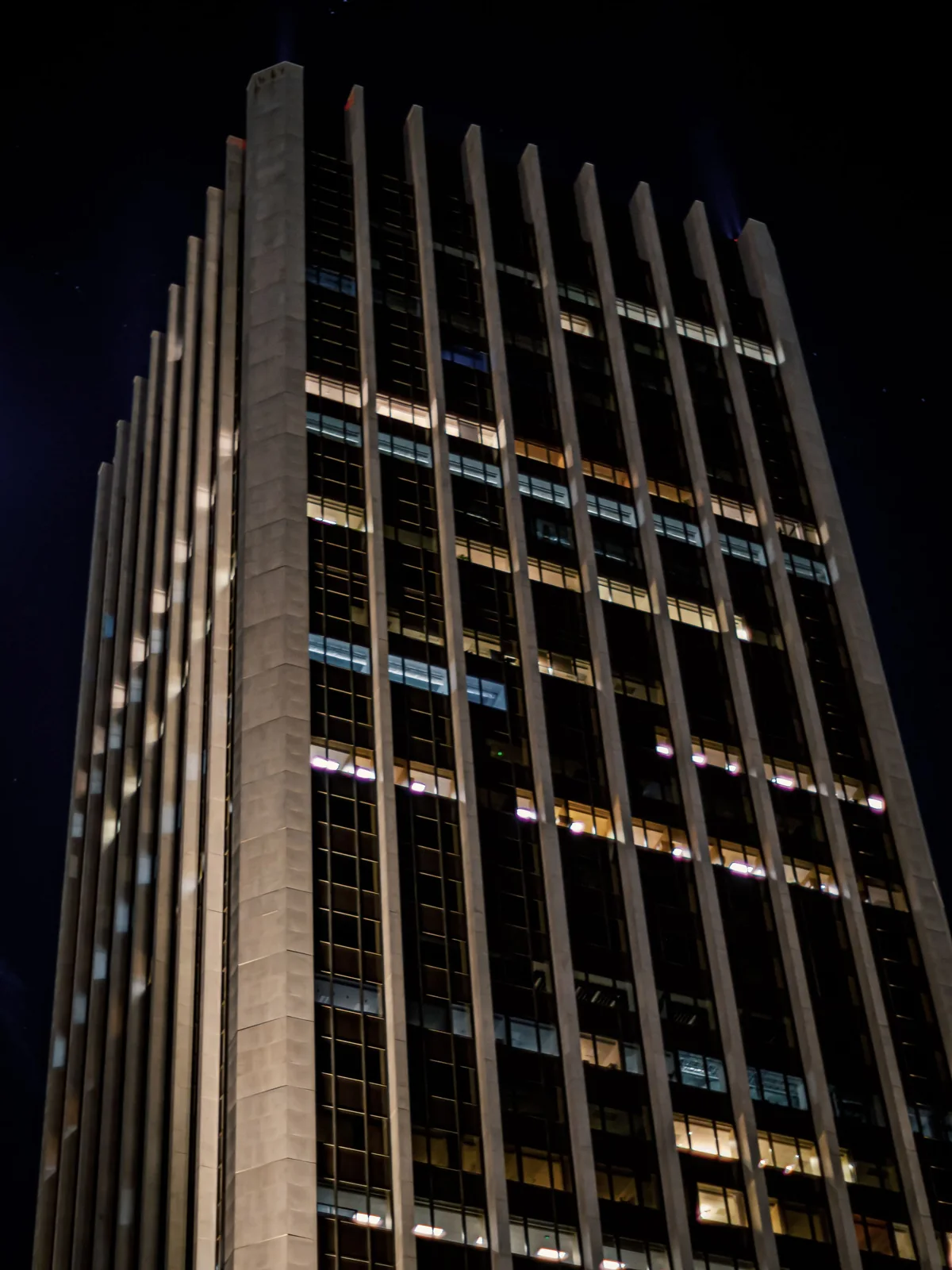 Contre-plongée nocturne sur une tour de bureaux aux cannelures de pierre, plusieurs plateaux allumés derrière le vitrage.