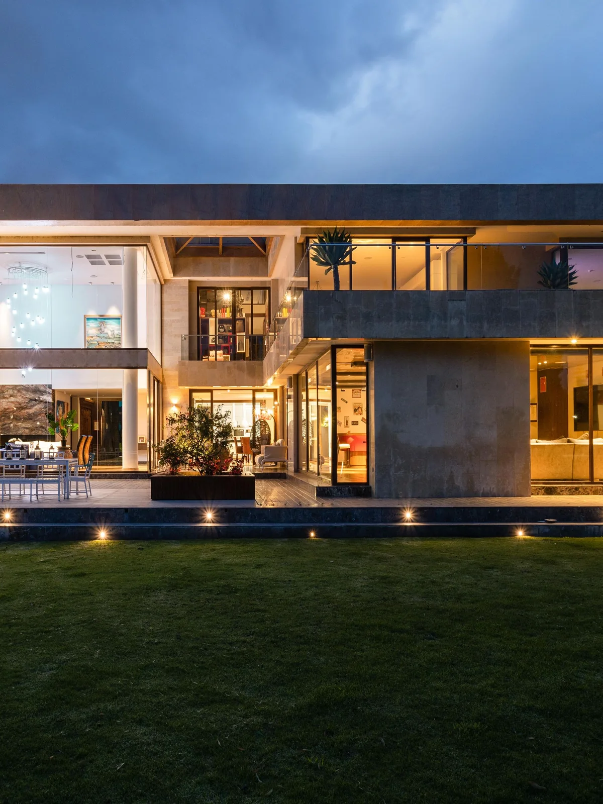 Villa contemporaine en pierre et verre à la tombée du jour, toutes ses baies allumées au-dessus d’une pelouse sombre.