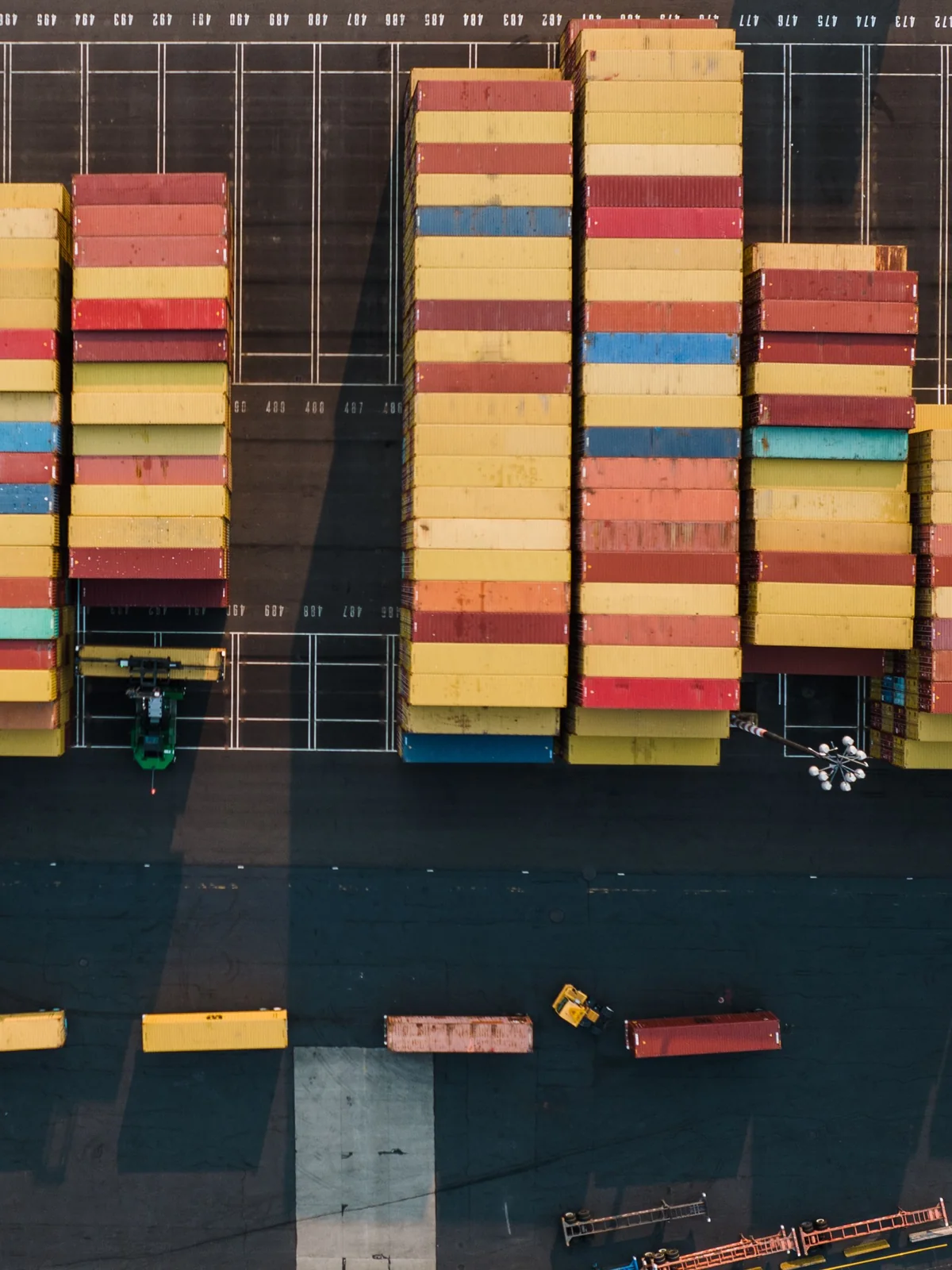 Vue verticale de drone sur une cour à conteneurs au soleil bas, des piles ocre et terre cuite dont les ombres s’étirent en diagonale sur le bitume.