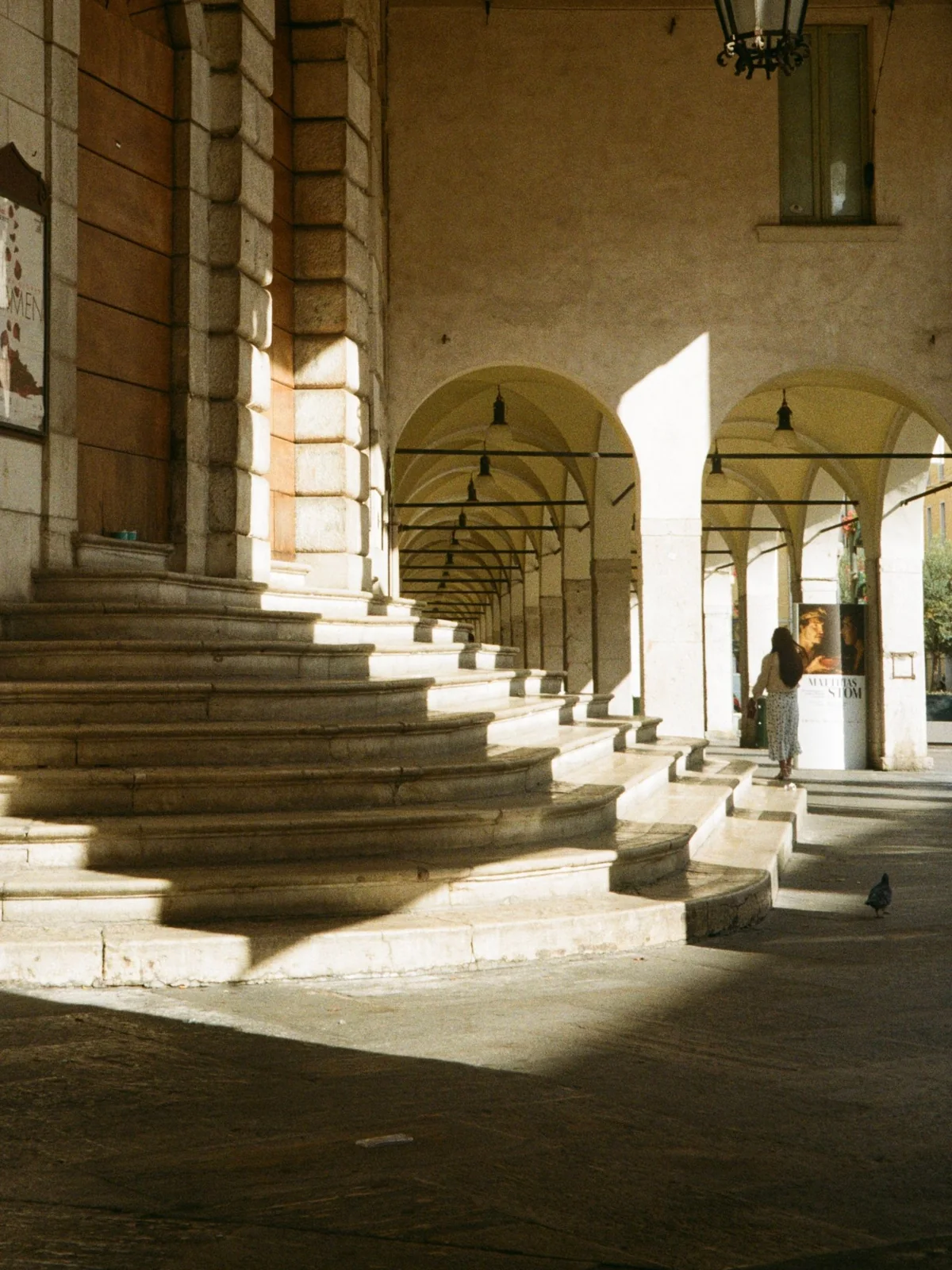 Galerie à arcades d’un bâtiment public du sud de l’Europe, un large escalier de pierre au premier plan et une silhouette qui marche au loin, dans la lumière rasante de fin de journée.