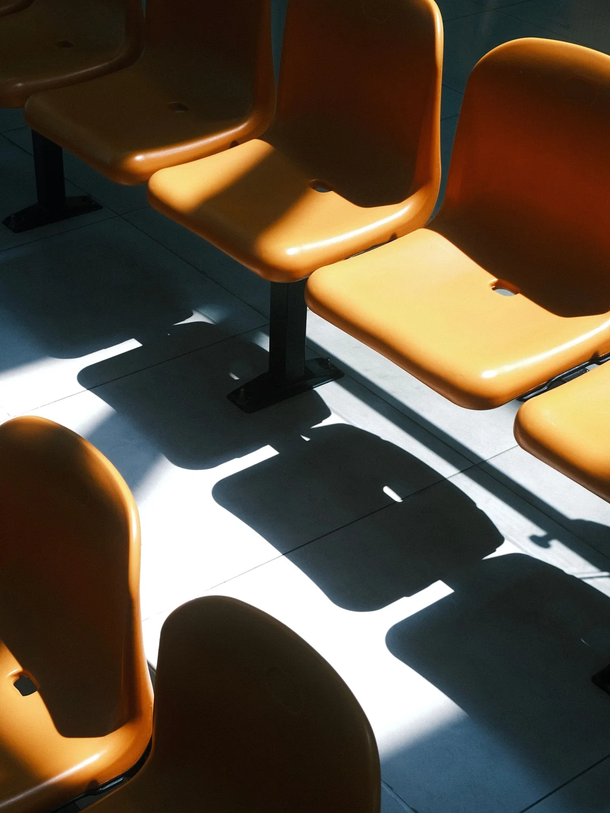 Une rangée de sièges en plastique ocre dans une salle d’attente, traversée par une bande de lumière dure qui projette leurs ombres nettes sur le carrelage gris bleuté.