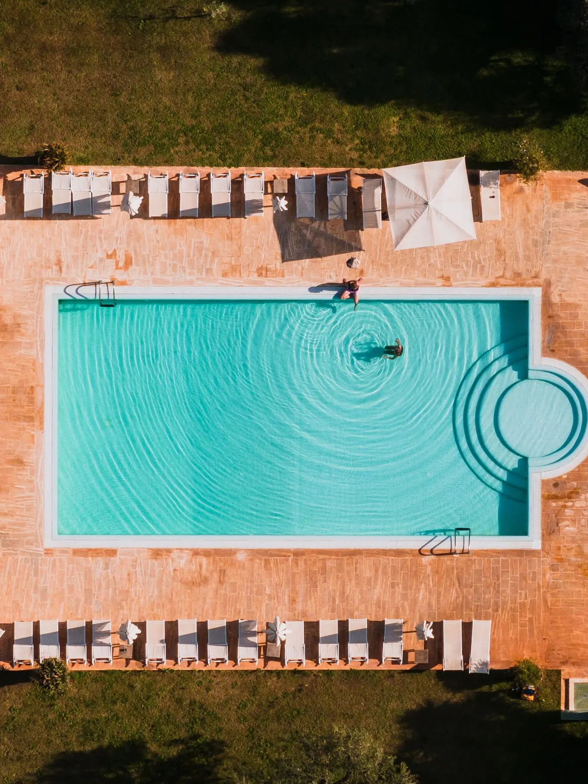 Vue aérienne à la verticale d’une piscine d’hôtel en Italie du Sud, dallage de terre cuite, deux rangées de transats blancs aux ombres allongées et un nageur seul au milieu de l’eau turquoise.